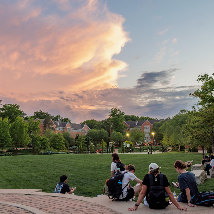 students sit on brick steps watching a pink sunset over campus